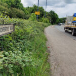 Shared use path with foliage to the left and a road name sign labelled "Coventry Road". To the right of the path is the main carriageway when a truck loaded with pipework has just gone past.