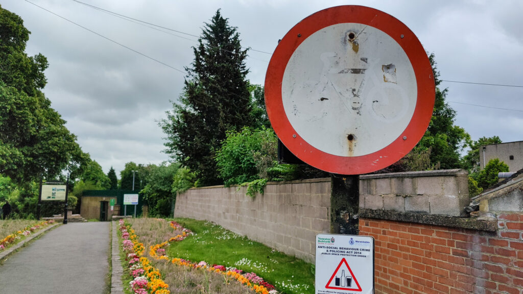 A worn "no cycling" sign at the entrance to Bedworth's Miners' Welfare Park. The sign is on a black post. A path leads into the picture from the left. Flower beds flank the path.